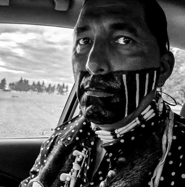 Image is a black and white photograph of a Blackfoot traditonal dancer with warpaint.